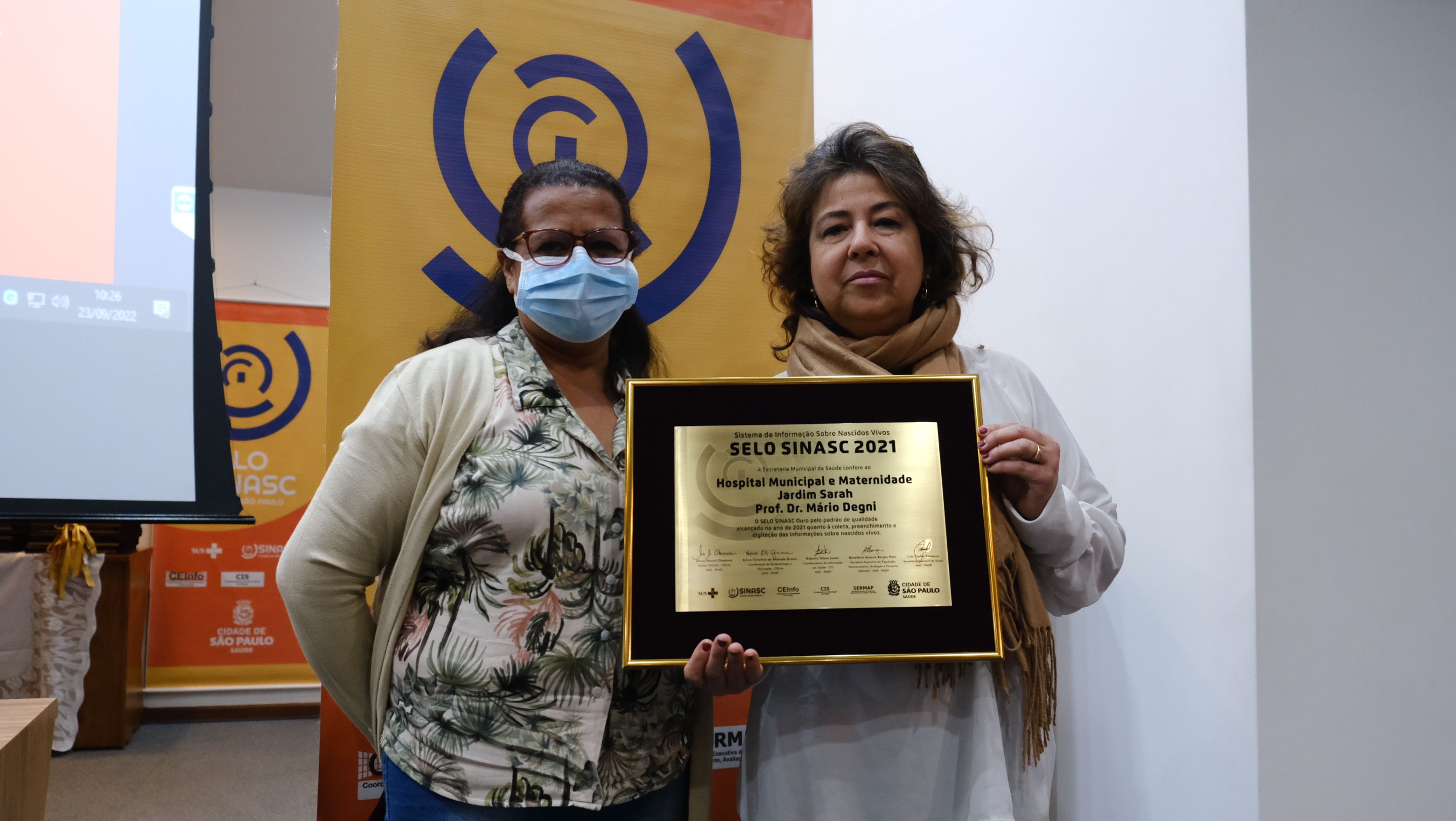 Na imagem há duas mulheres segurando uma placa de ouro. As duas estão em pé, uma delas usa óculos e uma camisa florida e a outra usa um cachecol marrom.